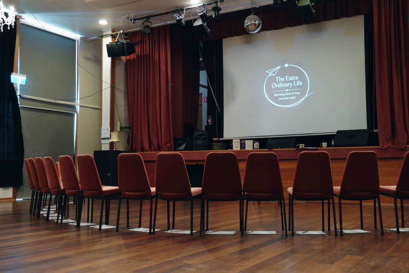An empty hall in Canberra set with a circle of chairs before a session begins, with the Extra Ordinary Life logo projected on the screen